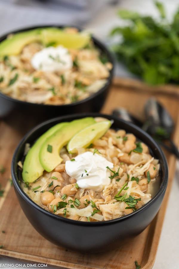 Close up image of two black bowls of white chicken chili with avocado and sour cream on a serving tray. 
