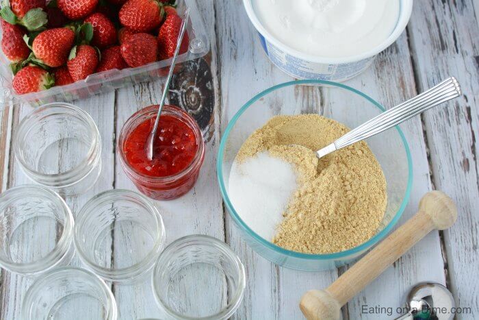 Ingredients needed for cheesecake - graham crackers, sugar, butter, cream cheese, whipped topping, strawberry preserves, fresh strawberries and fresh mint leaves and mason jars. 