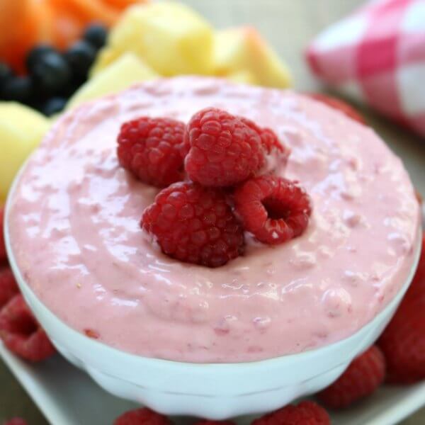 Close up of a bowl of raspberry fruit dip topped with fresh raspberries with more fresh fruit behind it for serving.  