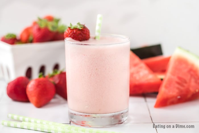 Close up image of a glass of watermelon strawberry smoothie with a strawberry and a straw in the glass. There is also pieces of watermelon and strawberries in the background. 