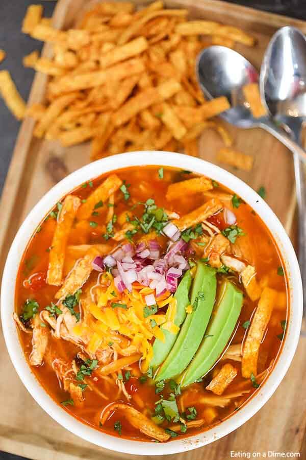 Close up image of a chicken tortilla soup in a white bowl with two silver spoons with tortilla strips in the background. 