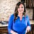 Close up, Carrie, a woman with long brown hair in a bright blue blouse, stands in a modern kitchen holding a kitchen utensil and smiling at the camera. The background features a granite countertop and stovetop.
