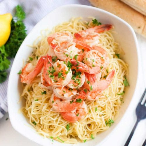 A white bowl filled with pasta topped with shrimp and chopped parsley, placed on a light surface. A lemon and some fresh herbs are partially visible in the background, suggesting a fresh and appetizing meal, related to a crock pot shrimp scampi recipe.