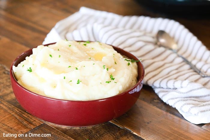 Close up image of mashed potatoes in a red bowl with a tea towel and a spoon on a table. 
