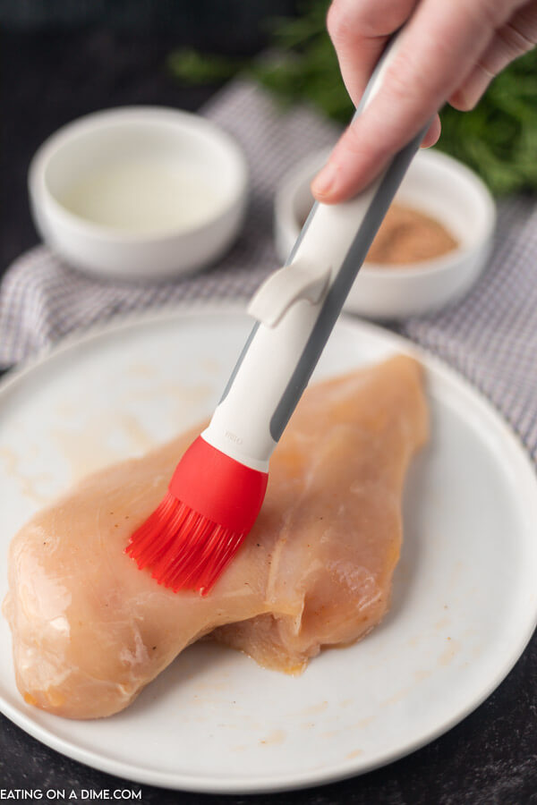 A photo showing the basting brush brushing the oil on a chicken breast on a white plate 
