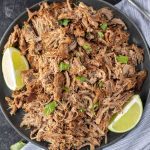 A plate of shredded, crispy slow cooker pork carnitas garnished with chopped cilantro, served with lime wedges on the side.