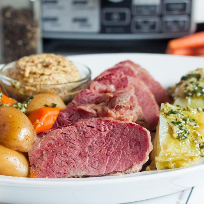 Close up of sliced corned beef on a white plate next to cabbage, carrots and potatoes with a small bowl of spicy mustard on the plate.  