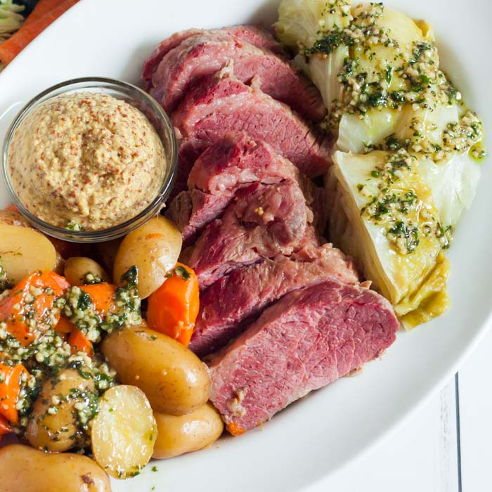 A large white platter with sliced corned beef in the center and cabbage, potatoes and carrots around it topped with garlic butter with a small bowl of grainy mustard on the plate as well.  
