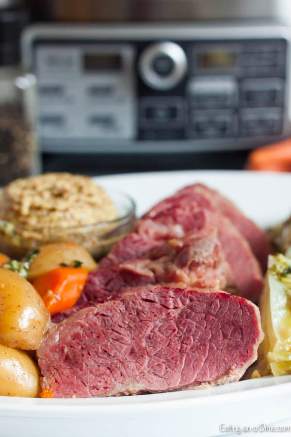 Sliced corned beef on a white plate next to cabbage, carrots and potatoes with a small bowl of spicy mustard on the plate.  