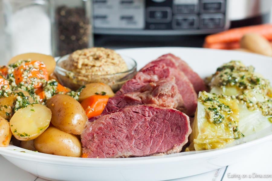 Sliced corned beef on a white plate next to cabbage, carrots and potatoes with a small bowl of spicy mustard on the plate.  The entire plate is sitting in front of a crock pot. 