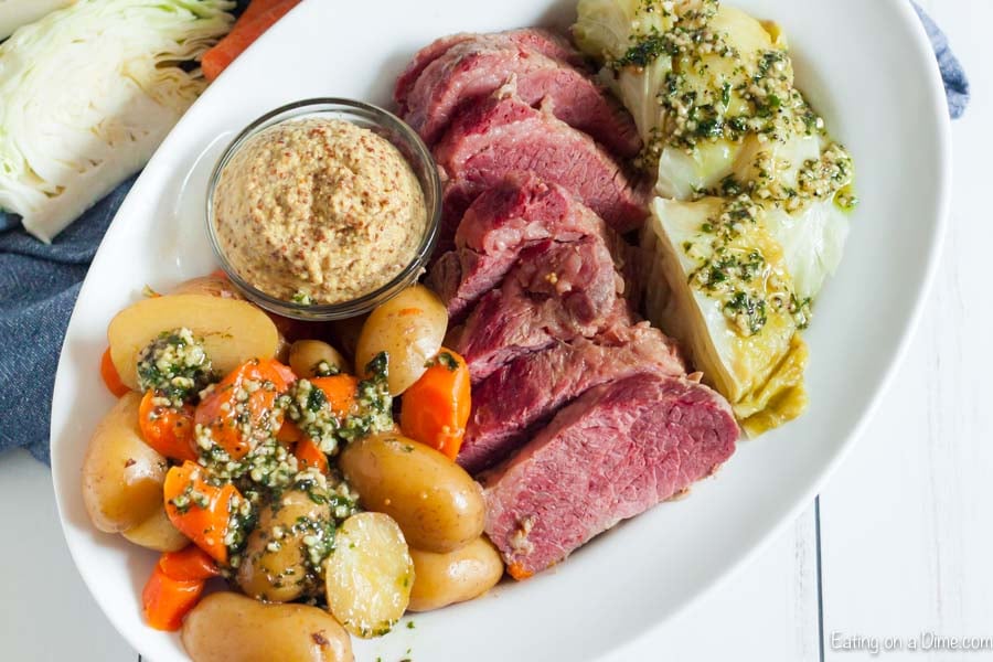 A large white platter with sliced corned beef in the center and cabbage, potatoes and carrots around it topped with garlic butter with a small bowl of grainy mustard on the plate as well.  