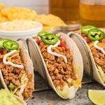 Three soft tacos filled with seasoned Crock Pot Ground Chicken Tacos, shredded lettuce, cheese, diced tomatoes, and topped with sour cream and jalapeño slices. Nacho chips and dip are blurred in the background.