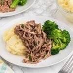 A white plate with mashed potatoes, crock pot shredded beef, and steamed broccoli is arranged neatly on a marble surface. A fork, bowl of mashed potatoes, and partial plate are visible in the background.