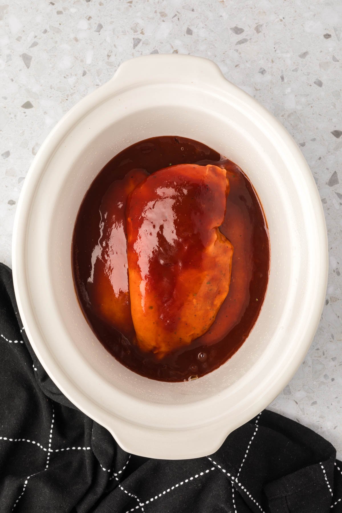Pouring BBQ Sauce over the top of seasoned chicken in the slow cooker
