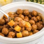 A white crock pot filled with Hawaiian meatballs and pineapple chunks in a brown sauce, with a metal serving spoon scooping some out.