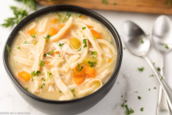 Close up image of chicken noodle soup in a black bowl with two silver spoons. 