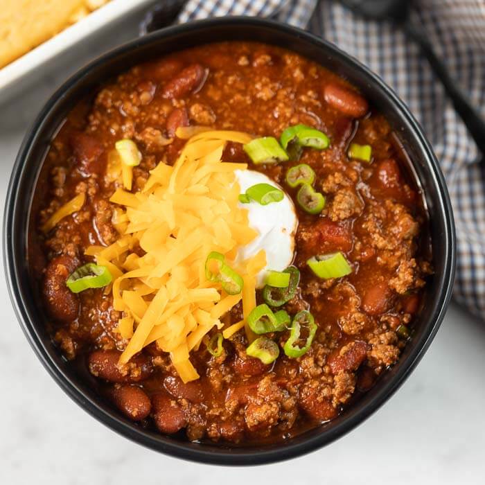 You are going to love this quick and easy Crockpot Chili Recipe - Try this easy slow cooker chili recipe that the entire family will love. This simple easy beef slow cooker chili recipe is the best! I love this easy simple homemade slow cooker chili recipe is delicious and simple to throw together in no time at all! #eatingonadime #recipesslowcooker #recipeseasy #recipesbest #slowcookerchilerecipeeasy #slowcookerrecipes #crockpotrecipes #chilirecipes Close up image of chili in a black bowl topped with cheese, sour cream and green onions.