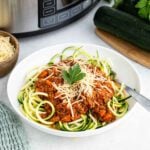 A white bowl of zucchini noodles topped with crock pot turkey bolognese, grated cheese, and a parsley garnish sits on a table near a slow cooker, fresh zucchinis, a bowl of shredded cheese, and a green cloth.