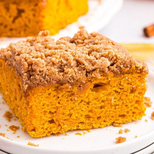 A close-up of a square slice from a pumpkin coffee cake recipe with a crumbly cinnamon streusel topping, served on a white plate. The cake appears moist and vibrant orange in color.