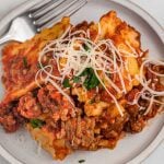 A plate of lasagna topped with shredded cheese and chopped parsley, showing layers of pasta, meat sauce, and melted cheese—reminiscent of crockpot ravioli and sausage—with a fork resting on the plate.