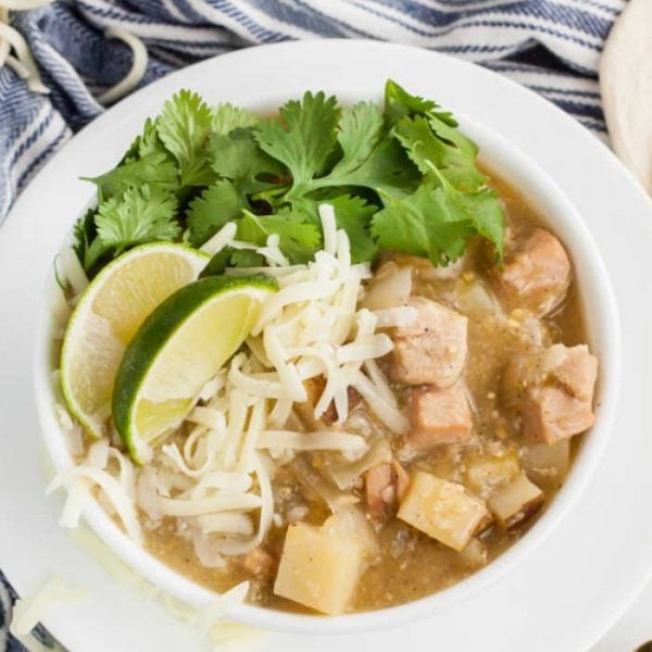 A bowl of Crock Pot Green Chili Stew with chunks of pork, potatoes, shredded cheese, lime wedges, and fresh cilantro, served on a white plate with a blue and white striped cloth underneath.