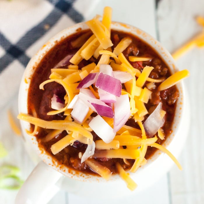 A bowl of cowboy chili topped with sour cream, cheese and red onions with 2 spoons next to it. 
