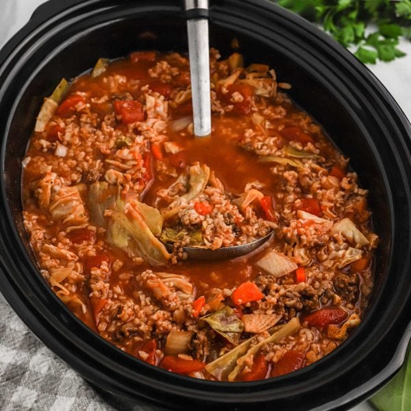 A Crockpot Cabbage Roll Soup simmers in the slow cooker, brimming with ground meat, rice, chopped cabbage, tomatoes, and carrots. A ladle rests inside while fresh parsley decorates the background.