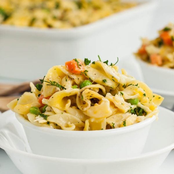 A white bowl filled with creamy chicken noodle casserole recipe, featuring wide egg noodles, peas, carrots, and shredded chicken, garnished with chopped parsley. Another bowl and a baking dish appear in the blurred background.