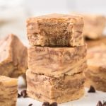 Three pieces of marbled chocolate peanut butter fudge are stacked on top of each other, surrounded by more fudge pieces and scattered chocolate chips on a white surface.