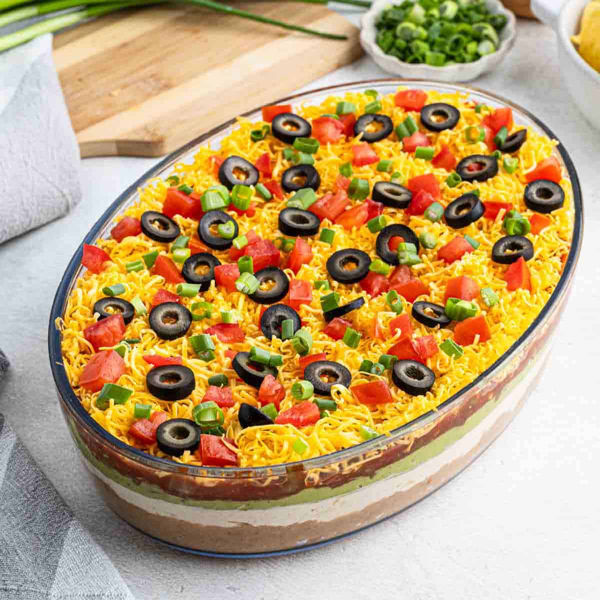 7 Layer Dip in a oval clear dish topped slice black olives, diced tomatoes, and chopped onions