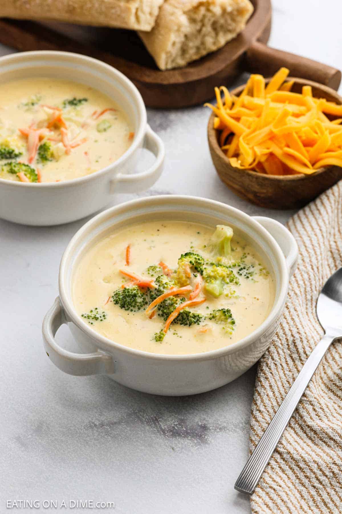 Two bowls of creamy panera broccoli cheddar soup garnished with shredded carrots and broccoli florets sit on a table beside a bowl of shredded cheddar cheese, a striped napkin, a spoon, and sliced bread on a wooden board.