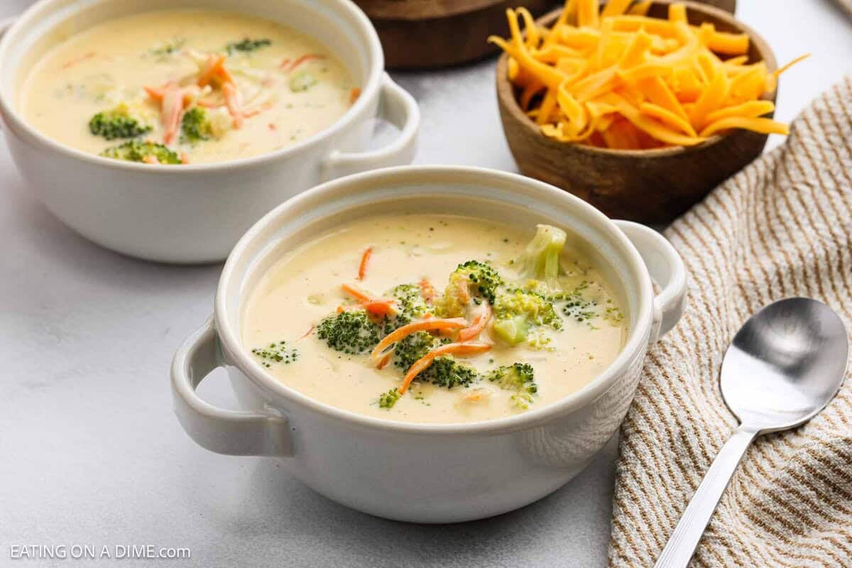 A bowl of creamy panera broccoli cheddar soup topped with broccoli florets and shredded carrots, with a spoon, striped napkin, and a bowl of shredded cheddar cheese nearby. Another bowl of this comforting soup sits in the background.