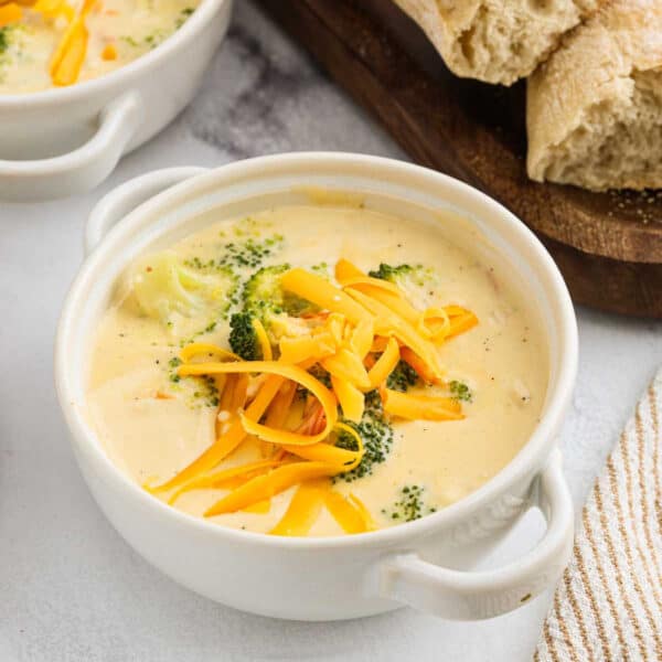 A white bowl filled with creamy panera broccoli cheddar soup, topped with shredded cheddar cheese. Pieces of broccoli are visible in the soup. Sliced bread is in the background.