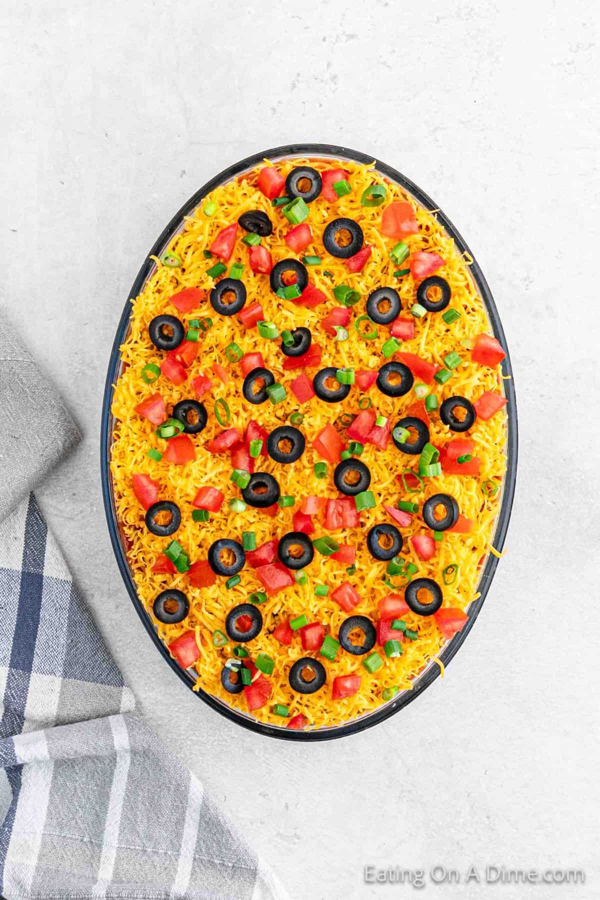 7 Layer Dip in a oval dish topped with slice black olives, diced tomatoes, and green onions