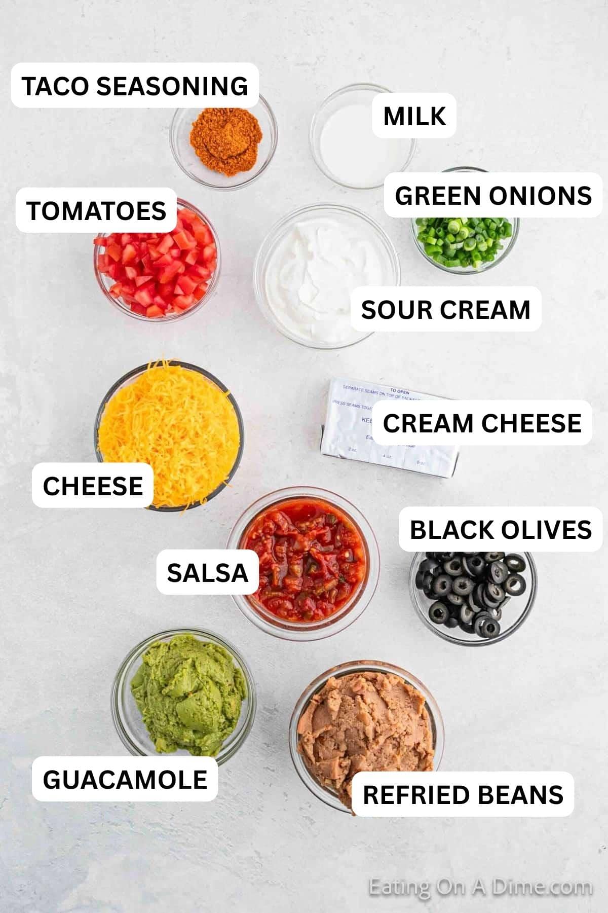 Ingredients needed to make this recipe which includes Salsa, refried beans, milk, cream cheese, sour cream, taco seasoning, guacamole, cheese, black olives, roma tomatoes, and green onions.