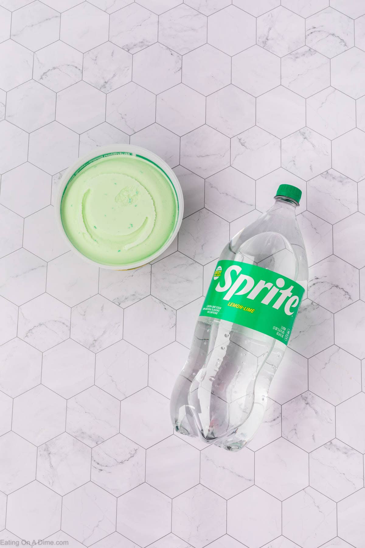 A tub of light green ice cream and a clear plastic bottle of Sprite lay on a white hexagon-tiled surface—perfect ingredients for a refreshing sherbet punch recipe.
