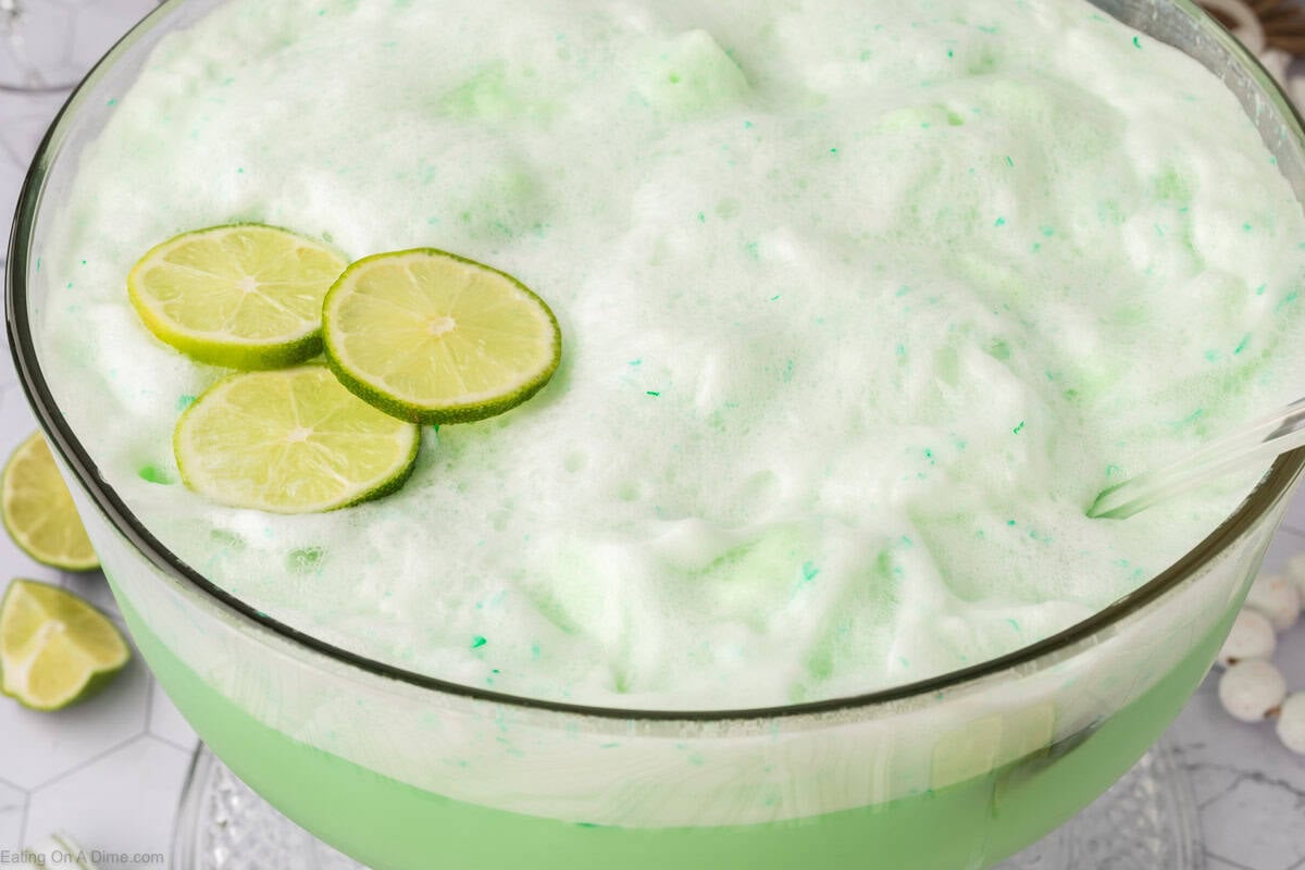 A large glass bowl filled with frothy, light green sherbet punch, topped with several slices of fresh lime. A serving spoon rests on the side of the bowl, ready to serve this classic sherbet punch recipe.