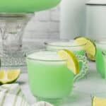 Three glass cups filled with light green sherbet punch, each garnished with a lime wedge. More lime slices and a green-and-white napkin are nearby, with a glass punch bowl rounding out this refreshing sherbet punch recipe scene.