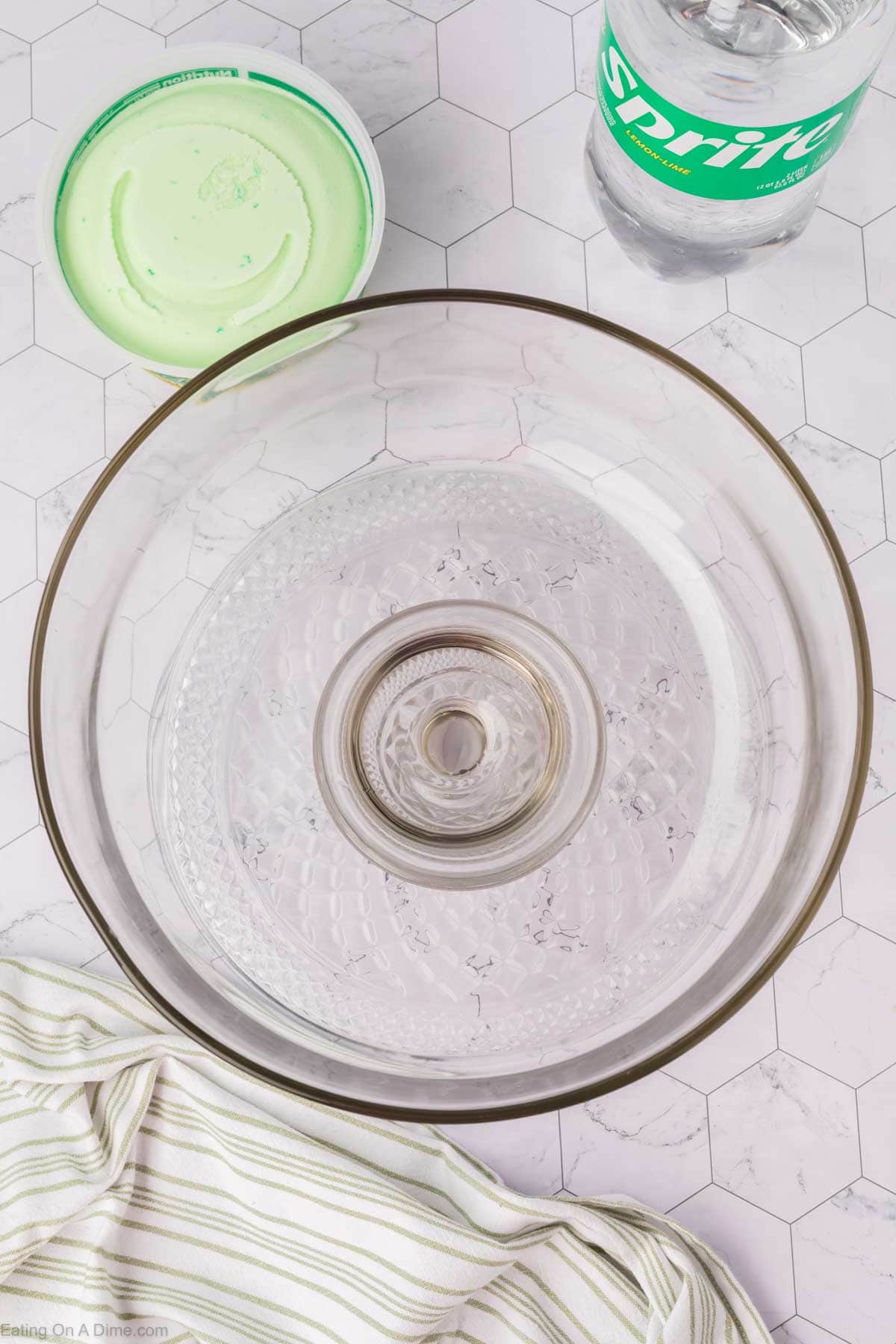 A large empty glass punch bowl sits on a white surface with a carton of green sherbet, a bottle of Sprite, and a striped kitchen towel nearby—perfect for preparing your favorite sherbet punch recipe.