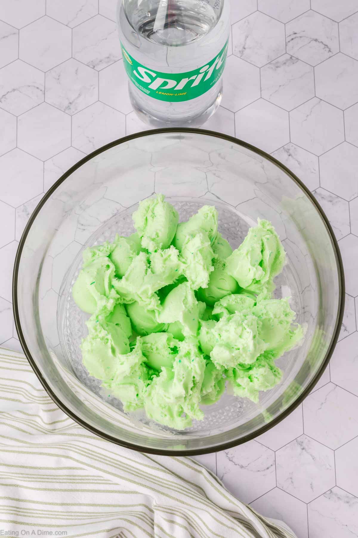 A glass bowl filled with scoops of light green sherbet sits on a hexagon-patterned surface next to a can of Sprite and a striped cloth napkin, ready for your favorite sherbet punch recipe.