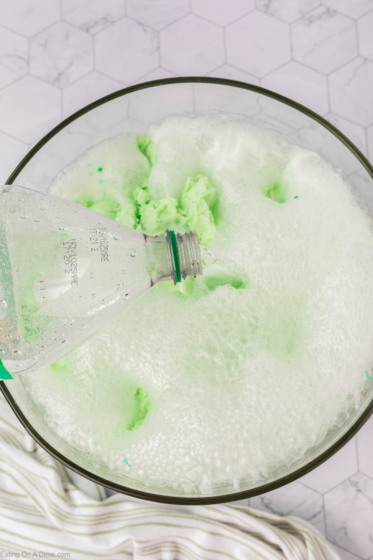 A close-up of a bowl filled with foamy, green bubbles as clear liquid is poured from a bottle, illustrating the refreshing start of a sherbet punch recipe against a white hexagonal tile background.