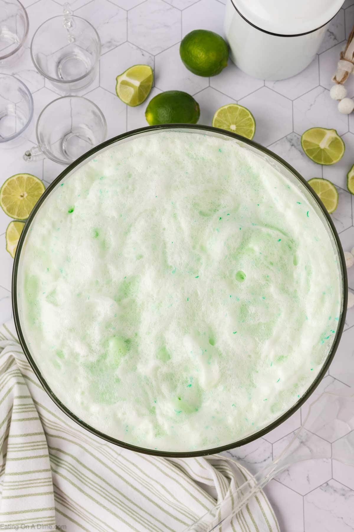 A large glass bowl filled with frothy, light green sherbet punch recipe sits on a striped cloth, surrounded by sliced and whole limes on a white hexagon tile surface. Punch glasses and a ladle are nearby.