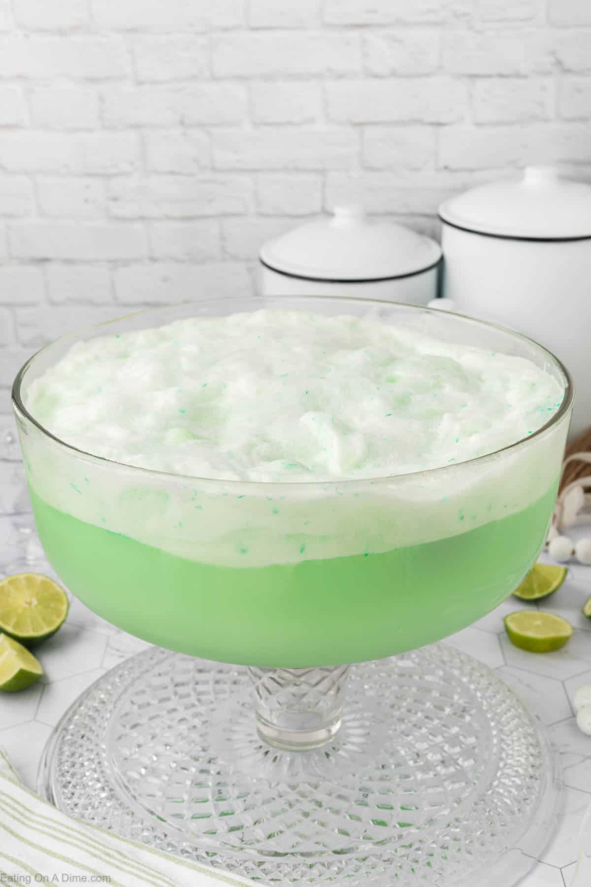 A glass punch bowl filled with a frothy, light green sherbet punch recipe sits on a clear platter. Fresh lime halves and white canisters are visible in the background on a white tiled surface.
