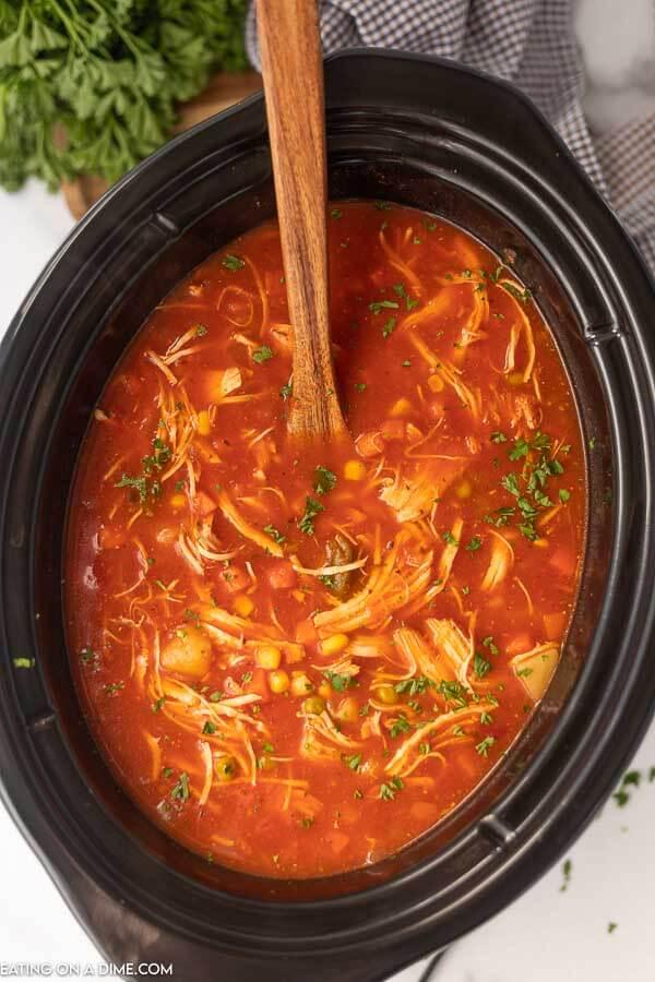 Overview of Chicken Vegetable Soup in a Black Crock Pot with a Wooden Spoon in it. 