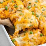 A close-up of easy chicken enchilada casserole, with cheesy enchiladas topped with chopped green onions and cilantro, as one enchilada is lifted from a baking dish using a wooden spatula.
