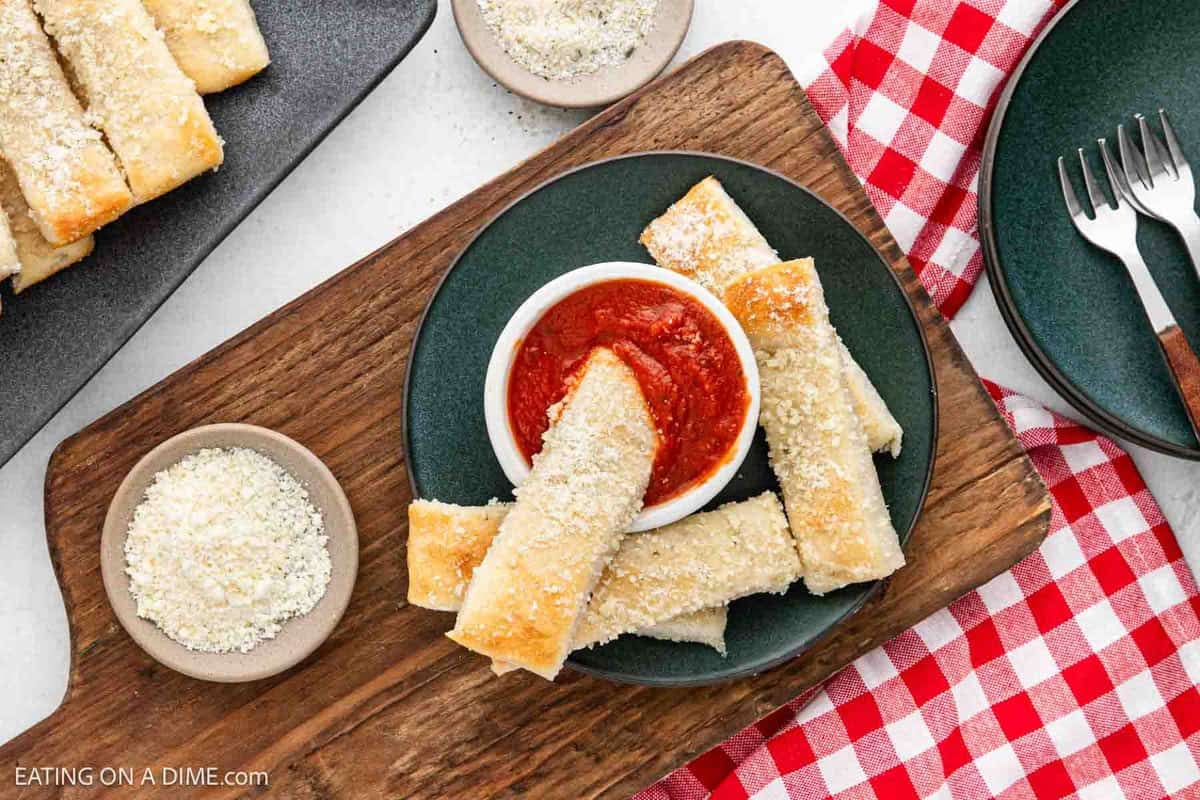 A top view of a wooden board showcasing Little Caesars Crazy Bread sprinkled with grated cheese around a small bowl of marinara sauce. The board rests on a table dressed in a red checkered cloth, accompanied by small bowls of grated cheese and two forks nearby.