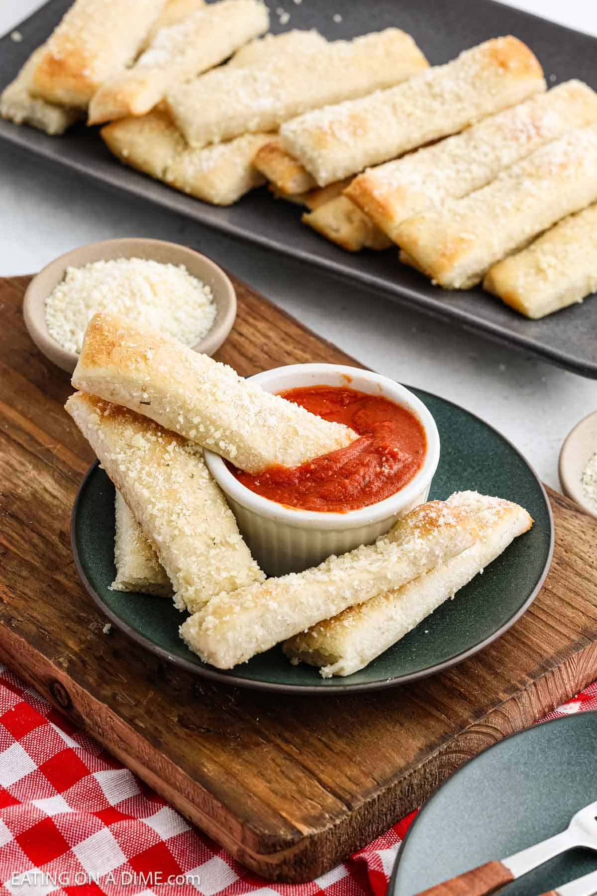 A plate of breadsticks, reminiscent of Little Caesars Crazy Bread, is stacked next to a small bowl of marinara sauce on a wooden board. More breadsticks are visible on a tray in the background, with a red-checked napkin peeking out at the bottom.