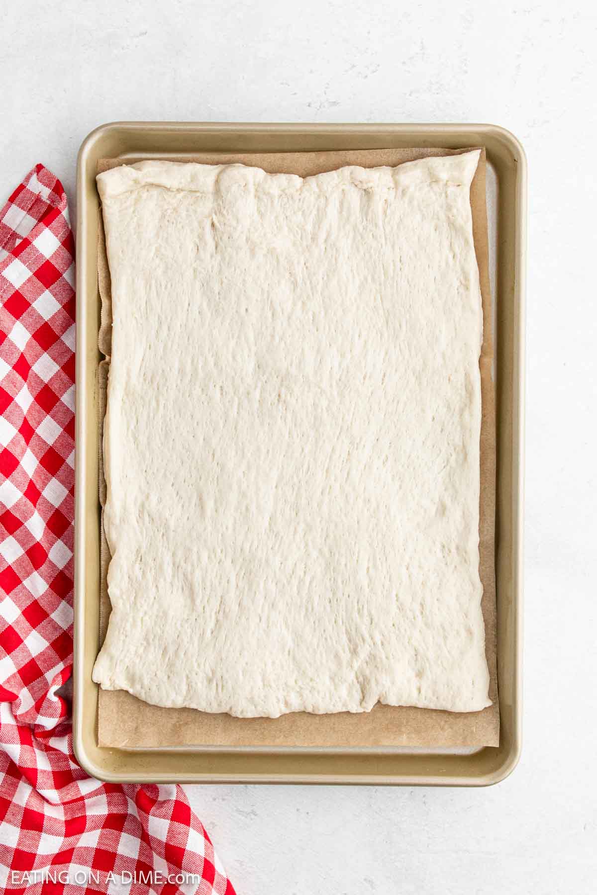 A sheet of rectangular pizza dough spreads on a parchment-lined baking tray, reminiscent of Little Caesars' Crazy Bread. A red and white checkered cloth rests on the left side of the tray, placed artfully on a pristine white surface.