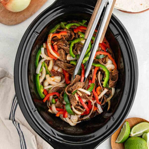A slow cooker filled with slow cooker fajitas—cooked beef strips, sliced green and red bell peppers, and onions. Metal tongs rest inside, with lime wedges and tortillas nearby on a white surface.