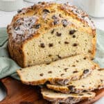 A round loaf of raisin bread, inspired by an Irish soda bread recipe, is partially sliced on a wooden board. Its golden crust and soft interior studded with raisins are displayed atop a green cloth.